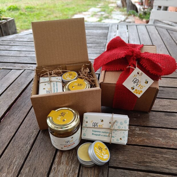 Confezione regalo (in esterno) aperta mostra i prodotti al suo interno, confezione chiusa con un fiocco rosso e in primo piano i tre prodotti: sapone, burro labbra e vasetto di miele.
